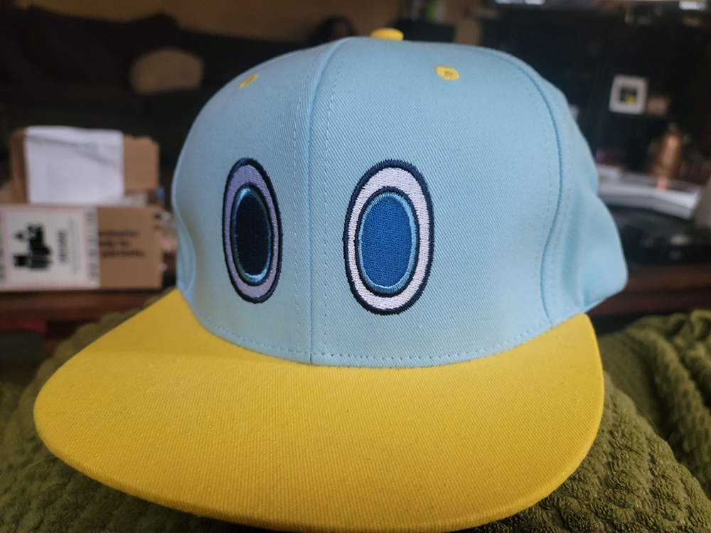 a snapback hat with embroidered eyes of a chao from sonic the hedgehog. its light blue with yellow brim and detailing